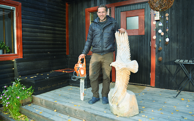Sebastian som står framför ett hus med en motorsåg i ena handen och en trä skulptur av en rovfågel i den andra.
